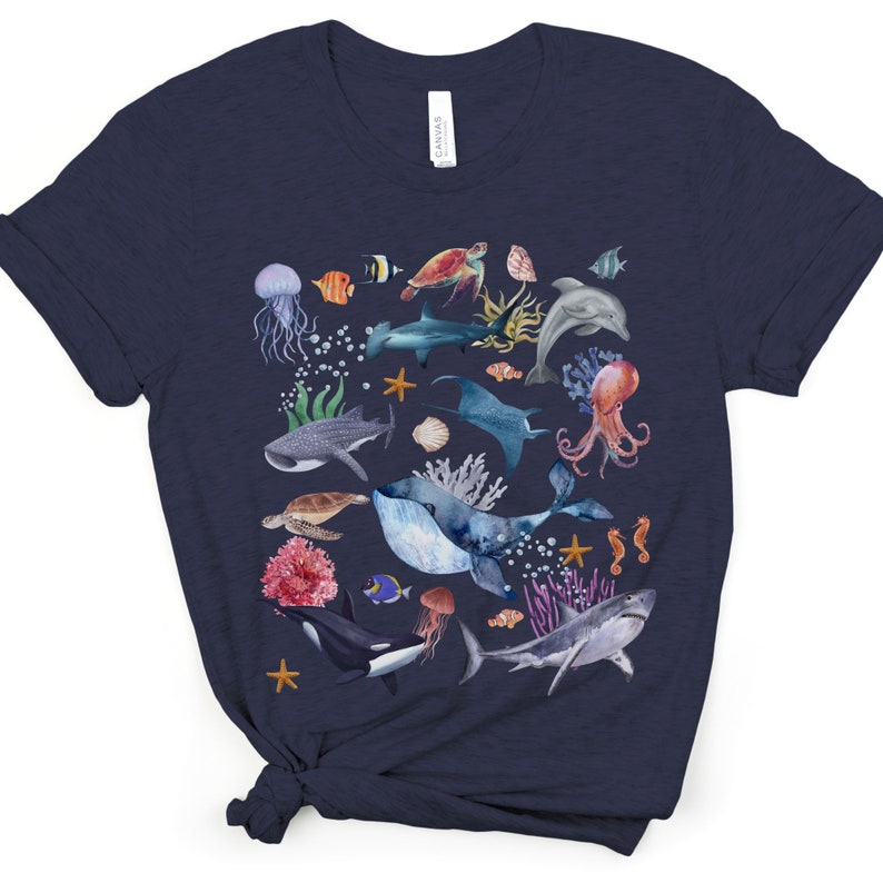 May include: A navy blue t-shirt with a watercolor design of various sea creatures, including a whale, a shark, a dolphin, a jellyfish, a seahorse, and coral.