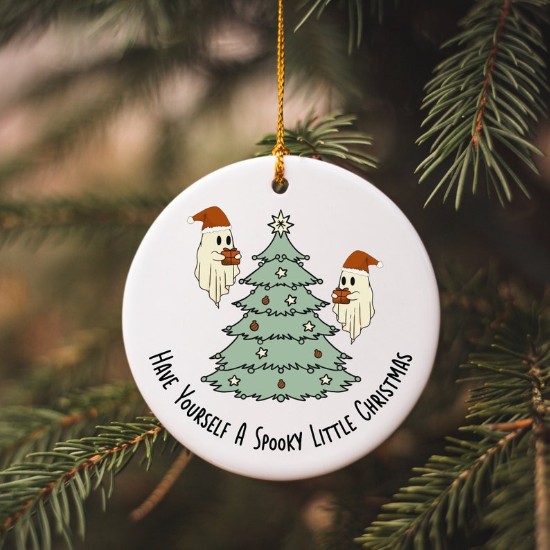 May include: White ceramic Christmas ornament with a gold string. The ornament features a green Christmas tree with star and ornament details, and two cartoon ghosts wearing Santa hats. Text reads "Have Yourself a Spooky Little Christmas."