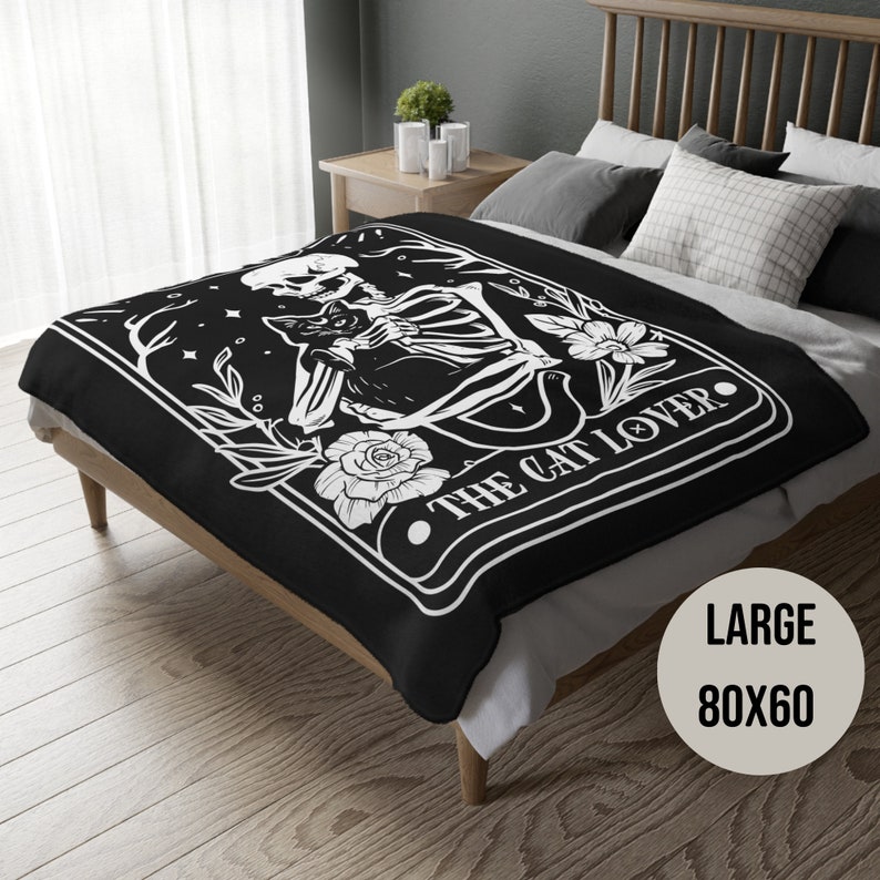 May include: Black fleece throw blanket with a white design of a skeleton holding a cat. The design is framed in a rectangle with the text "The Cat Lover". The blanket is on a bed with a white pillow and a black headboard. The text "Large 80x60" is in the bottom right corner of the image.