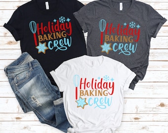 May include: Three t-shirts with the text "Holiday Baking Crew" in red, brown, and light blue. The design includes a whisk, snowflake, and gingerbread cookie. One black, one gray, and one white t-shirt are displayed with a pair of blue jeans.