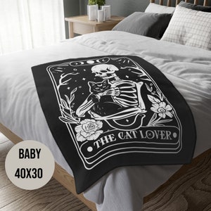 May include: Black and white throw blanket with a skeleton holding a black cat. The blanket features the text "The Cat Lover" in a decorative font.