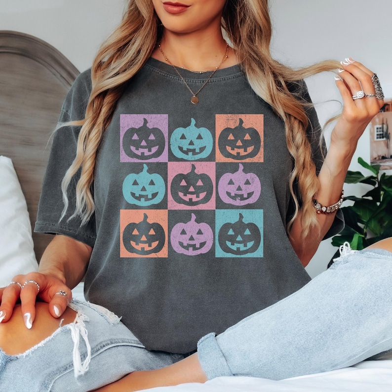 May include: Dark gray t-shirt featuring a grid of nine colorful jack-o'-lanterns. Each pumpkin is in a different color square: purple, blue, orange, and teal. The shirt is a comfortable, casual style.