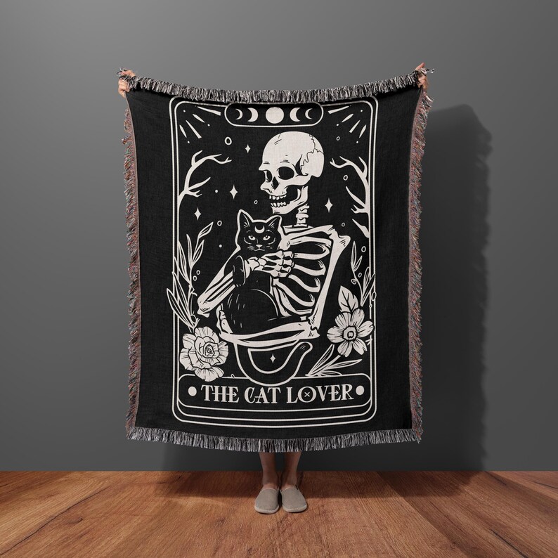 Puede incluir: Tapiz tejido en blanco y negro con un esqueleto sosteniendo un gato negro. El tapiz tiene un dise&ntilde;o de carta de tarot con el texto "THE CAT LOVER" en la parte inferior.