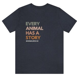 May include: Dark navy blue t-shirt with the text "EVERY ANIMAL HAS A STORY" in green, tan, and orange. The hashtag "#ANIMALRESCUE" is below the text. The shirt is a crew neck and has short sleeves.