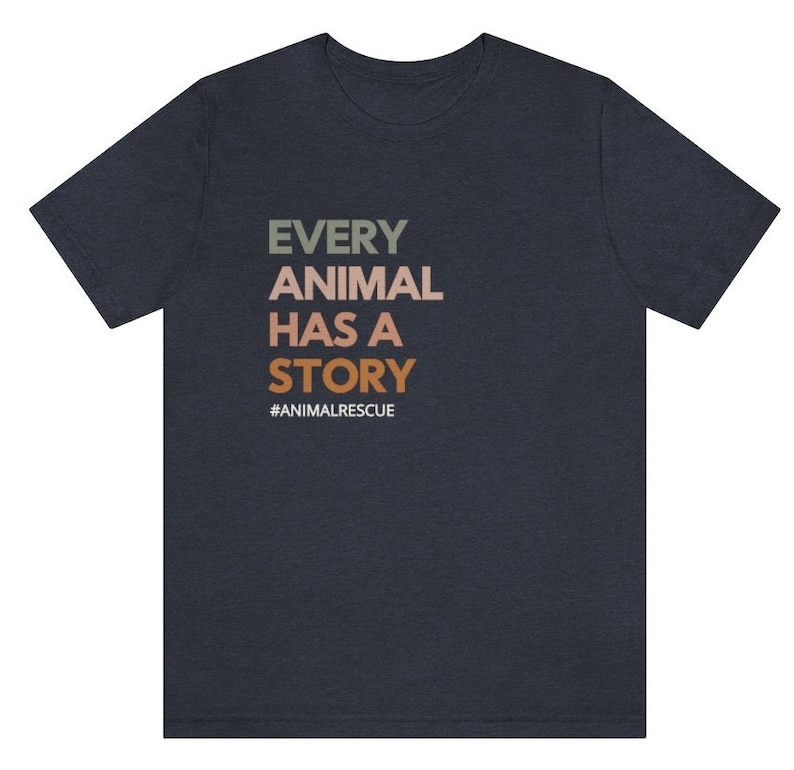 May include: Dark navy blue t-shirt with the text "EVERY ANIMAL HAS A STORY" in green, tan, and orange. The hashtag "#ANIMALRESCUE" is below the text. The shirt is a crew neck and has short sleeves.