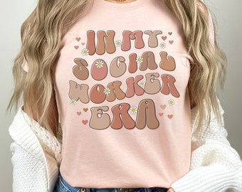 May include: A pink t-shirt with the text "In My Social Worker Era" in a retro font with brown and peach accents. The text is surrounded by small white daisies and pink hearts.