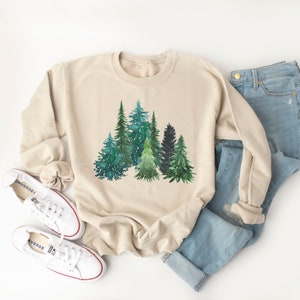 May include: A beige sweatshirt with a watercolor design of a forest of green trees. The sweatshirt is folded and laid flat on a white surface.