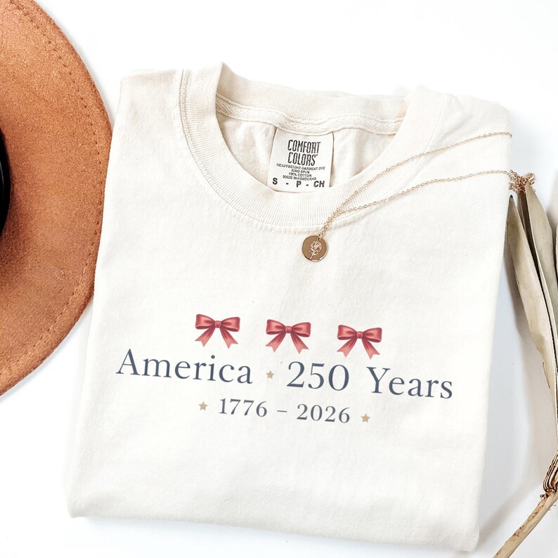 May include: Cream-colored t-shirt with the text "America 250 Years" and the years "1776-2026" printed in dark blue. The design includes three red bows. A gold necklace with a pendant is draped across the shirt.