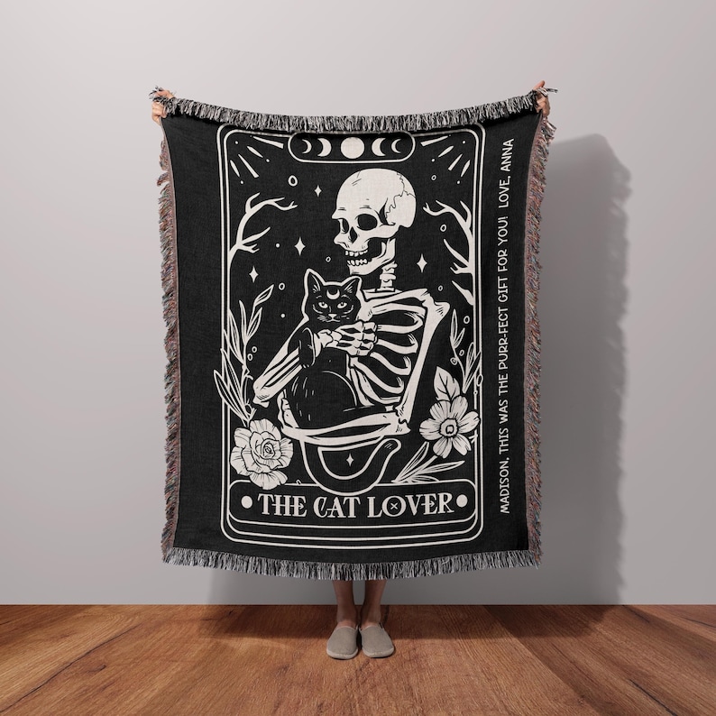 Puede incluir: Tapiz tejido en blanco y negro con un esqueleto sosteniendo un gato negro. El tapiz tiene un dise&ntilde;o de carta de tarot con las palabras "THE CAT LOVER" y "MADISON, THIS WAS THE PURR-FECT GIFT FOR YOU! LOVE, ANNA".