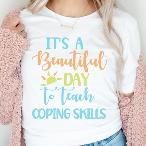 Pu&ograve; includere: T-shirt bianca con la scritta "It's a Beautiful Day to Teach Coping Skills" in blu, arancione e verde. La maglietta ha una grafica a forma di sole e viene indossata con un cardigan rosa e jeans blu.