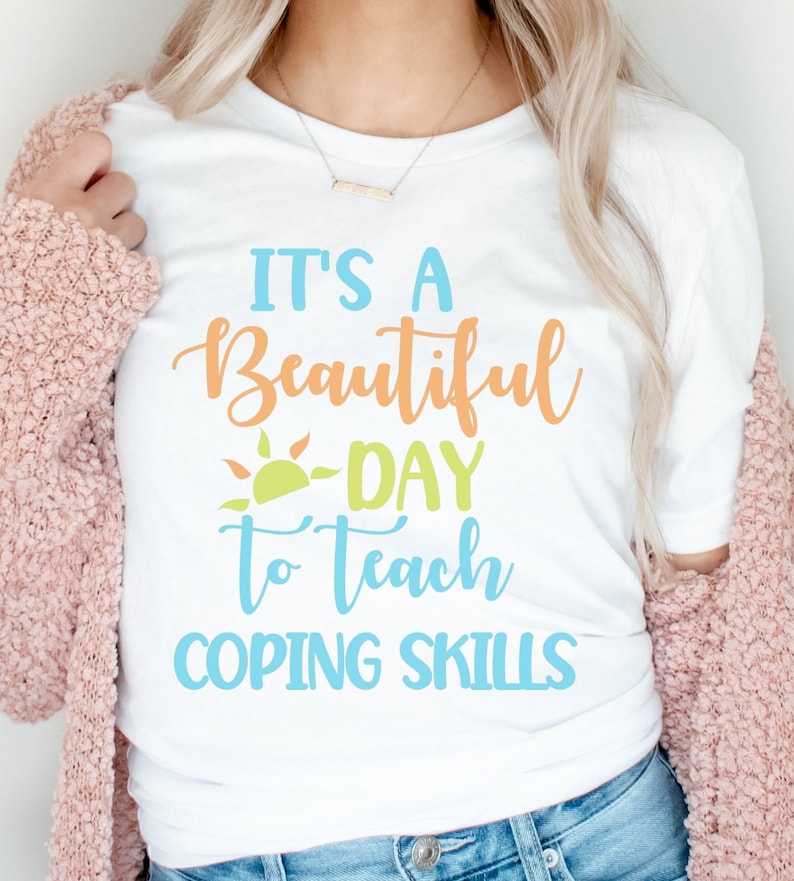 Pu&ograve; includere: T-shirt bianca con la scritta "It's a Beautiful Day to Teach Coping Skills" in blu, arancione e verde. La maglietta ha una grafica a forma di sole e viene indossata con un cardigan rosa e jeans blu.