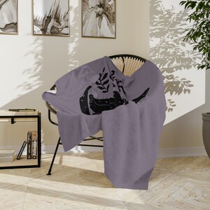 May include: A soft, plush throw blanket in a muted purple colour with a black and white design of a hand holding a branch with leaves. The blanket is draped over a wicker chair.