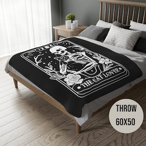 May include: Black and white throw blanket featuring a skeleton holding a cat with the text "The Cat Lover". The blanket is 60 inches by 50 inches.