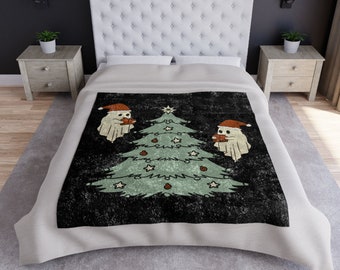 May include: A Christmas-themed blanket featuring a distressed black background with a light green Christmas tree decorated with stars and ornaments. Two ghost figures wearing Santa hats are on either side of the tree. The blanket is on a bed with a white headboard and nightstands.