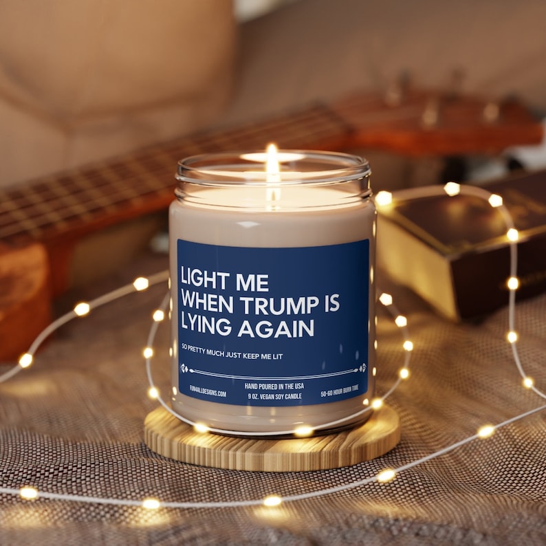 Puede incluir: Una vela de soja azul y blanca con el texto "Enci&eacute;ndeme cuando Trump mienta de nuevo. As&iacute; que, b&aacute;sicamente, mantenme encendida." La vela est&aacute; sobre un posavasos de madera y rodeada de luces.