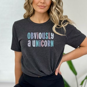 Op de afbeelding: Een donkergrijs T-shirt met de tekst "OBVIOUSLY a UNICORN" in pastelkleuren.
