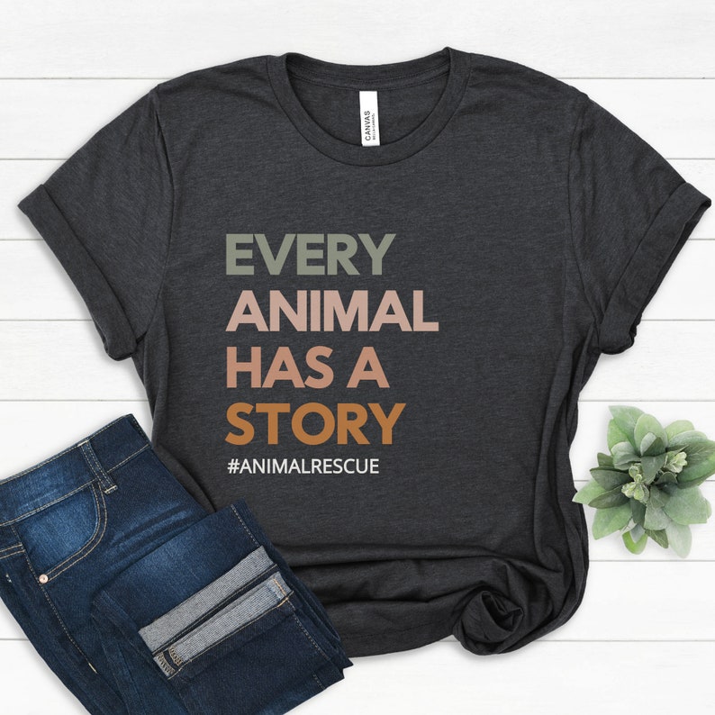 May include: Dark gray t-shirt with the text "EVERY ANIMAL HAS A STORY" in green, pink, and orange. The hashtag "#ANIMALRESCUE" is below the text. The shirt is paired with blue jeans.