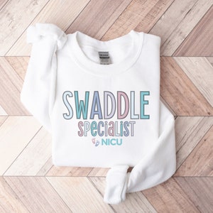 May include: White sweatshirt with the text "SWADDLE specialist NICU" in pastel colors. The text is outlined in a darker shade of the same color. There are two small footprints below the text.