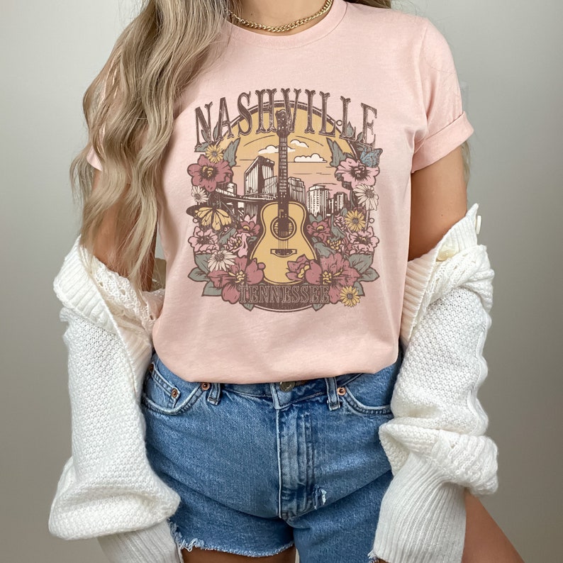Pu&ograve; includere: Una maglietta rosa con una grafica in stile vintage di una chitarra, fiori e il testo "Nashville Tennessee".
