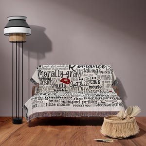 May include: A white throw blanket with black text and a red lip print. The text includes words like "romance", "morally gray", "gothic", "stalking haunts", "dominance", "cat &", "filthy", "little mouse", "escaped prison", "obsession", and "secrets".