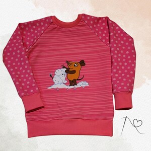 Könnte beinhalten: Ein pinkfarbenes Langarm-Sweatshirt mit einem Cartoon-Maus- und Schneemann-Motiv. Die Ärmel haben ein weißes Schneeflockenmuster. Der Körper des Shirts hat horizontale Streifen.