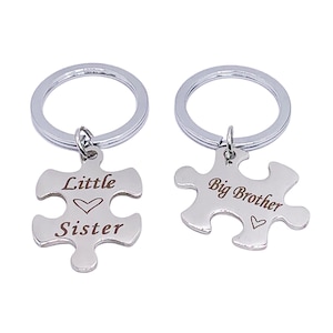 May include: Two silver keyrings with puzzle pieces attached. The left keyring has the words "Little Sister" and a heart on the puzzle piece. The right keyring has the words "Big Brother" and a heart on the puzzle piece.