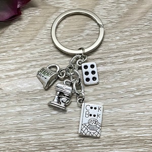 Op de afbeelding: Zilverkleurige sleutelhanger met bak-thema bedels. De bedels tonen een maatbeker, een keukenmixer, een domino met zes stippen en een kookboekbedel met het woord "Cookbook" en een cupcake.