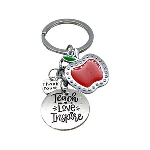 Könnte beinhalten: Ein silberner Schlüsselanhänger mit einem roten Apfel-Anhänger, einem silbernen Anhänger mit dem Text "Teach Love Inspire" und einem kleinen silbernen Herz-Anhänger mit dem Text "Thank You".