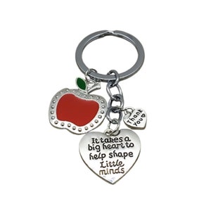 Peut inclure: Porte-clés argenté avec un charme de pomme rouge, un charme de cœur avec le texte "Il faut un grand cœur pour aider à façonner les petits esprits", et un petit charme de cœur avec le texte "Merci".