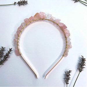 Puede incluir: Una diadema dorada con cristales de cuarzo rosa alambrados a su alrededor. La diadema está hecha de un metal delgado y flexible y está diseñada para ajustarse cómodamente a la cabeza.