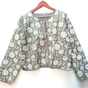 May include: A green and white floral print jacket with a quilted design. The jacket has a round neckline, long sleeves, and two front pockets. The interior lining is a black and white striped pattern. The jacket is hanging on a wooden hanger.