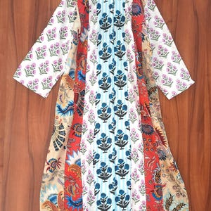 May include: A long-sleeved, patchwork dress with a mandarin collar. The dress features panels of various floral patterns in white, red, blue, and beige. The sleeves and parts of the dress have a white background with purple floral prints. The dress is hanging on a wooden hanger.