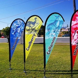 May include: Four teardrop-shaped flags with black borders stand on a grassy area. Each flag features a different design and color: blue with a koala, yellow with an emu, teal with a kangaroo, and red with a stylized image. The flags have text: Nargoon, Coolberry, Callanna, and Wanbi.