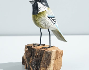 May include: A hand-painted wooden bird figurine featuring a black head with white markings, a yellow-tinted chest, and blue-toned wings. The bird stands on thin black metal legs attached to a natural, rough-cut wooden block base.