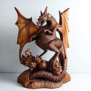 May include: A hand-carved wooden sculpture of a dragon with detailed scales, large wings, and sharp horns. The creature is depicted standing on a textured, rocky base with its tail coiled around the bottom, showcasing intricate wood grain patterns.