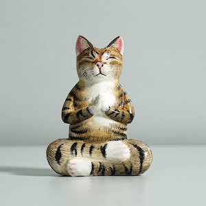 May include: A ceramic figurine of a tabby cat sitting in a yoga pose with its paws folded in front of its chest. The cat has closed eyes and a serene expression.