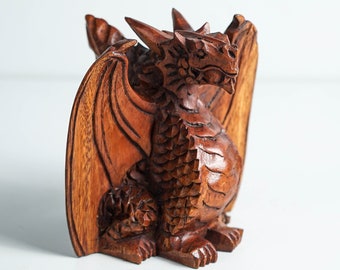 May include: A hand-carved wooden dragon figurine with intricate details. The dragon is brown and has its wings spread out. It is sitting on a white surface.
