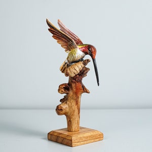 May include: A hand-carved wooden hummingbird sculpture with red, green, and white painted details, perched on a natural wood branch attached to a square wooden base. The bird has an elongated black beak and wings extended in a flight position.