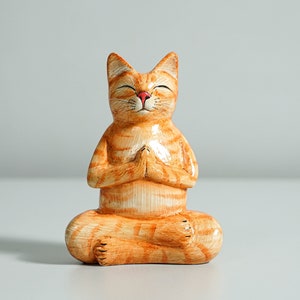 May include: Orange and white ceramic cat figurine sitting in a yoga pose with its eyes closed. The cat has a red nose and is meditating.