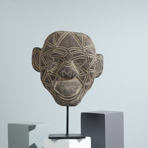May include: A wooden sculpture of a face with geometric patterns carved into it. The sculpture is brown and black and is mounted on a black stand.