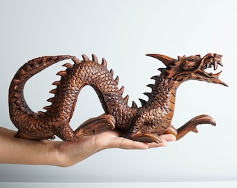 May include: A hand holds a dark brown wooden dragon sculpture. The dragon is curled in a defensive position with its head raised and mouth open.