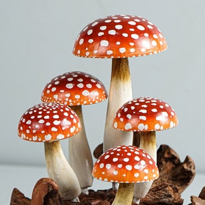 May include: A cluster of five decorative mushrooms with vibrant red caps dotted with white spots. The stems are a creamy white color, and the mushrooms are attached to a piece of natural wood. The overall aesthetic is whimsical and nature-inspired.