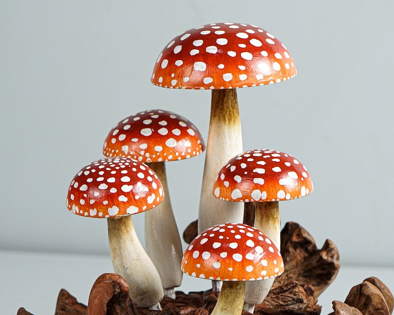 May include: A cluster of five decorative mushrooms with vibrant red caps dotted with white spots. The stems are a creamy white color, and the mushrooms are attached to a piece of natural wood. The overall aesthetic is whimsical and nature-inspired.