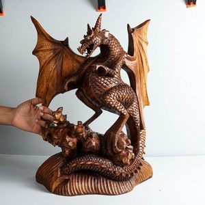 May include: A hand-carved wooden dragon sculpture featuring intricate scale detailing, large spread wings, and a long tail coiled around a rocky base. The creature is depicted in a standing position with its head turned and mouth open.