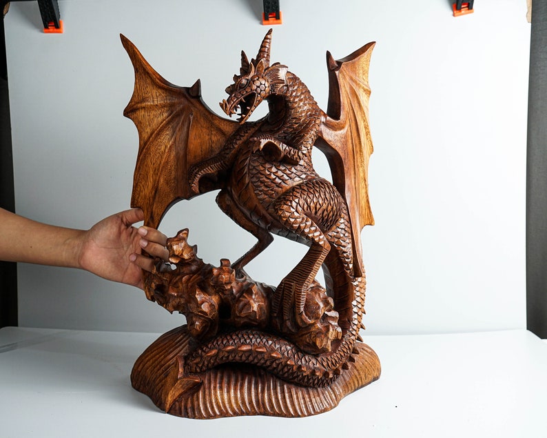 May include: A hand-carved wooden dragon sculpture featuring intricate scale detailing, large spread wings, and a long tail coiled around a rocky base. The creature is depicted in a standing position with its head turned and mouth open.