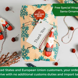 May include: A rectangular package wrapped in tan paper with a teal snowflake pattern, tied with red and white twine. A circular red Wood Glass Decor sticker, a white Thank You tag, and a small wooden Santa Claus ornament are attached.