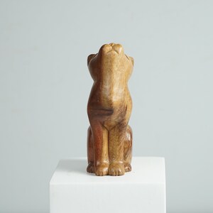 May include: A hand-carved wooden figurine of a cat in a seated position with its head tilted upward. The sculpture features natural wood grain patterns with varying shades of light tan, medium brown, and deep reddish-brown tones throughout the body.