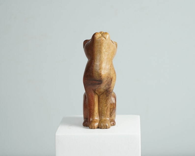May include: A hand-carved wooden figurine of a cat in a seated position with its head tilted upward. The sculpture features natural wood grain patterns with varying shades of light tan, medium brown, and deep reddish-brown tones throughout the body.