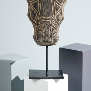 May include: A wooden sculpture of a stylized face with geometric patterns, mounted on a black metal stand. The sculpture is brown and has a weathered finish.