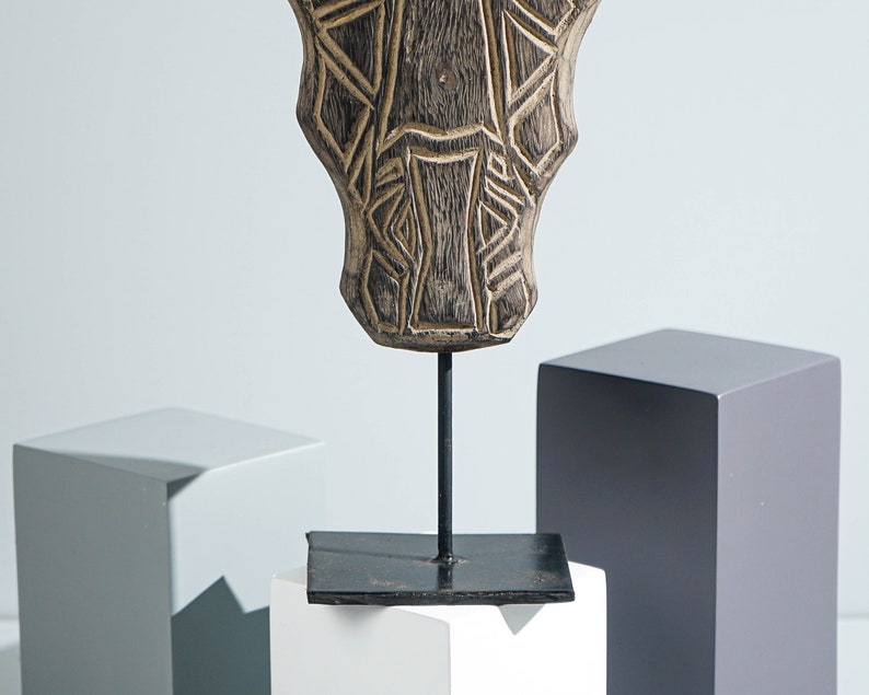 May include: A wooden sculpture of a stylized face with geometric patterns, mounted on a black metal stand. The sculpture is brown and has a weathered finish.